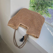 Woven straw tote bag with tassel, resting on windowsill, front view