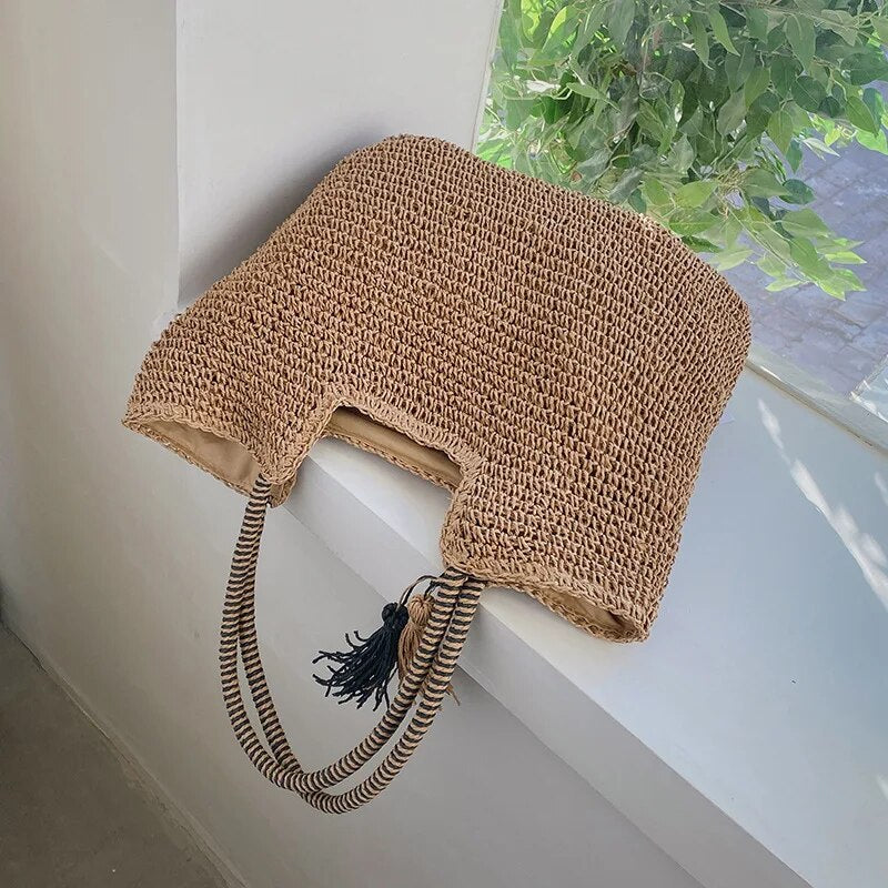 Woven straw tote bag with tassel, resting on windowsill, front view